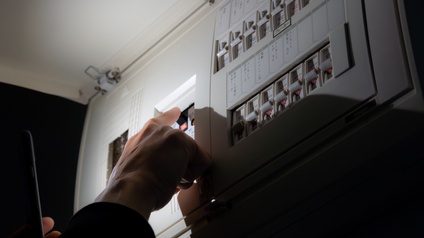 Nahaufnahme der Hand einer Person, die bei einem geöffneten Stromkasten den Fi-Schutzschalter drückt