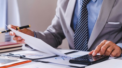 Nahaufnahme einer hinter einem Tisch sitzenden Person, die ihren linken Zeigefinger auf einem Taschenrechner hat. Die rechte Hand hält ein Papier und einen Stift. Auf dem Tisch liegen mit Statistiken und Diagrammen bedruckte Zettel