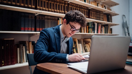 Eine Person sitzt in einer Bibliothek an einem Tisch voller Bücher, einem Laptop und lernt. Im Hintergrund stehen unzählige Bücher in Bücherregalen.