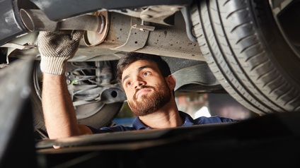 Der Kopf einer Person sowie die rechte Hand mit Handschuh sind unter einem Auto. Die Person blickt auf ein langes Rohr und hält dieses mit der rechten Hand. Links und rechts von der Person sind Reifen