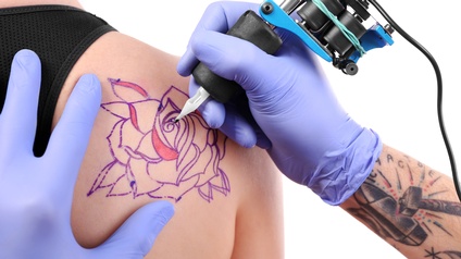 Nahaufnahme von der Erstellung eines Tattoos auf der Schulter einer Person mit einer Tätowiermaschine und lila Handschuhen
