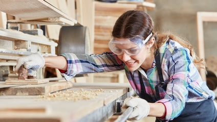 Person mit Ohrenschutz und Schutzbrillen hält Holzbalken auf Hobelbank, ringsum Holzspäne liegend