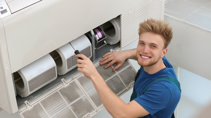 Lächelnde Person mit Bart blickt hinauf während sie mit Schraubenzieher in Klimaanlage schraubt