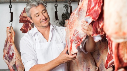 Person in weißer Schürze betrachtet großes Fleischstück, das zum Trocken aufgehängt ist, im Hintergrund an Haken weitere Fleischstücke