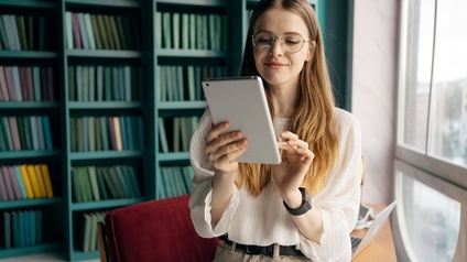 Jugendliche Person mit langen Haaren, Brille und weißer Bluse hält ein Tablet in Händen, im Hintergrund steht eine Couch sowie Regale mit Büchern vor einer offenen hellen Fensterfront