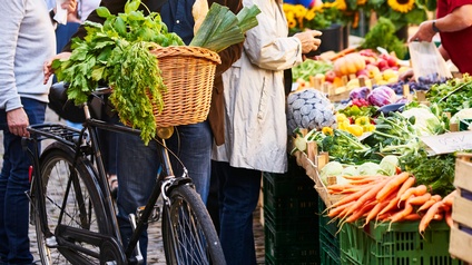 Mehrere Personen stehen um Markstand mit Gemüse, daneben ein Fahrrad mit Gemüse im Korb
