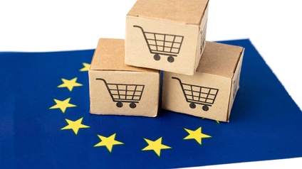 Drei kleine Kartonagen mit Aufdruck eines Einkaufswagen-Symbols aufeinander gestapelt auf kleiner EU-Fahne platziert: blauer Stoff mit gelben in Kreisform angeordneten Sternen