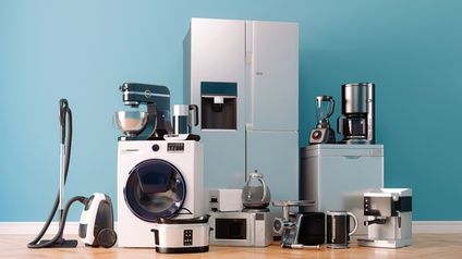 Vor einer blauen Wand auf einem Holzboden sind eine Waschmaschine, ein Staubsauger, ein Kühlschrank, eine Mikrowelle, Wasserkocher, ein Fleischwolf, zwei unterschiedliche Mixer, zwei Kaffeemaschinen, ein Toaster und weitere Haushaltsgeräte