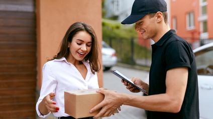Zwei Personen stehen sich im Freien neben einem Gebäude gegenüber. Die rechte Person übergibt der linken Person ein kleines Paket und hält ein Klemmbrett. Die linke Person freut sich