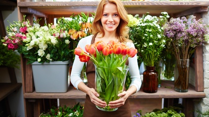 Lächelnde Person mit Schürze hält Vase mit orangen Tulpen in Händen vor Regal mit verschiedenen Schnittblumen stehend