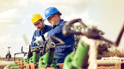 Personen mit Arbeitskleidung, Schutzhelme und Schutzbrillen halten einen Laptop in den Händen und blicken darauf während sie bei ei Gasrohren stehen
