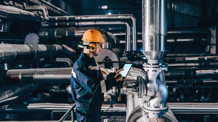 Person in Arbeitskleidung mit orangem Helm und Gehörschutz in Seitenansicht steht in Industrieanlage vor großen Metallrohren und hält Tablet in Händen