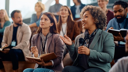 Mehrere Personen in Bürokleidung sitzen in einem Auditorium und blicken nach vorne. Die Person rechts unten im Bild hält in der linken Hand ein Mikrofon