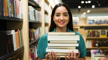 Junge Frau steht mit einem Stapel Bücher im Arm in einer Buchhandlung