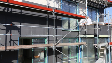 Hausfassade mit sichtbaren schwarzen Dämmplatten umgeben von Baugerüst, eine Person auf Gerüst in Rückenansicht beim Anbringen von Dämmplatten auf Fassade