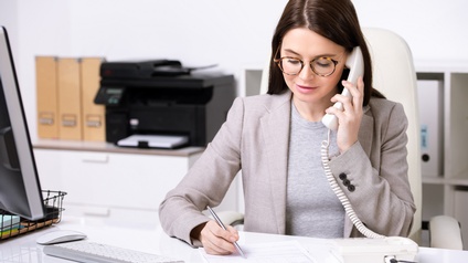 Eine Person sitzt an einem Schreibtisch und hält den Hörer eines Festnetztelefons ans Ohr. Gleichzeitig schreibt sie mit einem Stift auf einem Blatt Papier. Auf dem Tisch sind unter anderem ein Bildschirm, eine Tastatur und eine Maus