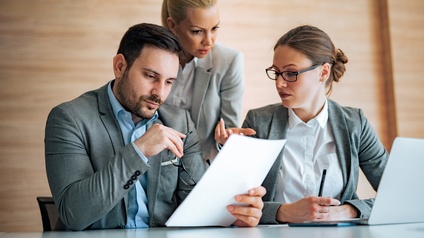 Zwei Personen in Bürokleidung sitzen nebeneinander an einem Tisch. Hinter ihnen steht eine weitere Person. Die links sitzende Person hält einen Zettel. Alle drei blicken auf den Zettel