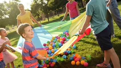 Erwachsene Personen spielen mit einer Gruppe von Kindern im Freien auf einer Wiese. Sie halten alle eine Ecke eines bunten Fallschirms in der Hand, auf der kleine, bunte Bälle liegen, die balanciert werden müssen