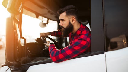 Person mit dunklen Haaren und Bart und rot-kariertem Hemd sitzt in einem Führerhaus eines LKWs und blickt  in den Rückspiegel.