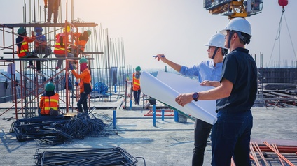 Zwei Personen mit Helmen stehen direkt nebeneinander auf einer Baustelle. Die linke Person hält einen Plan. Die rechte Person deutet in die Ferne. Im Hintergrund bauen Personen ein kleines Gerüst zusammen