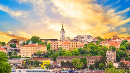 Panoramaaufnahme der Stadt Belgrad mit historischen und modernen Gebäuden. In der Mitte ragt ein Kirchturm in die Höhe. Im Vordergrund ist ein Fluss mit Booten. Am Himmel sind Wolken