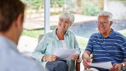 Im linken Bildrand ist die Rückenansicht einer Person. Ihr gegenüber sitzen zwei Personen mit kurzen, weißen Haaren und Brillen. Beide halten jeweils ein paar Zettel in den Händen. Hinter ihnen ist ein Garten