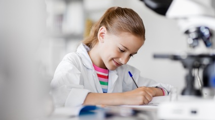 Lächelndes Kind in weißem Kittel schreibt mit Kugelschreiber und blickt darauf, im Vordergrund verschwommen Mikroskop
