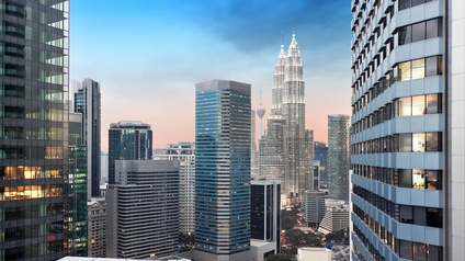 Aufnahme vieler moderner Wolkenkratzer, die unterschiedlich hoch sind. Die Fassaden bestehen teilweise aus großen Gläserfronten. Im Hintergrund ist ein Turm. Teilweise brennen in den Wolkenkratzer Lichter