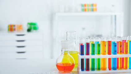 Zwei Laborgläser mit oranger und gelber Flüssigkeiten, nebenstehend Phiolen mit bunten Flüssigkeiten, im Vordergrund Glaspipette und Trägerglas,