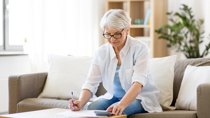 Person mittleren Alters mit Brille sitzt auf einer Couch in einem Wohnraum und stützt sich auf einen Beistelltisch ab während sie dabei auf Unterlagen blickt und einen Stift sowie einen Taschenrechner bedient