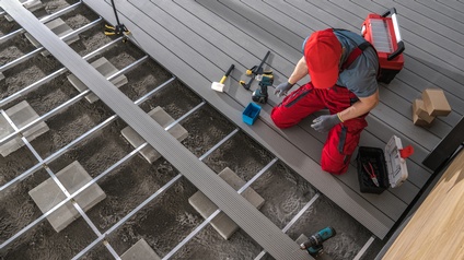 Eine Person in roter Latzhose, mit Handschuhen und mit roter Kappe kniet auf den Brettern einer Terrasse. Um die Person liegen verschiedene Werkzeuge. Vor der Person ist der Terrassenboden nicht fertig gebaut