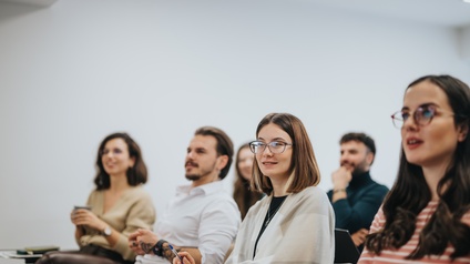 Seitliche Aufnahme von sechs Personen, die in zwei Reihen hintereinander in einem Raum sitzen und alle in dieselbe Richtung blicken