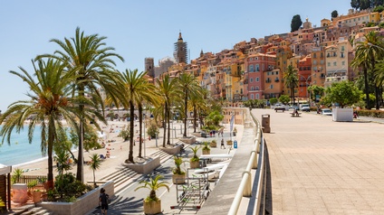 Eine Küstenstadt mit dicht gebauten, farbigen Häusern an einem Hang. Im Vordergrund ist eine breite Promenade mit Geländer, daneben ein Strand mit einzelnen Personen. Das Meer liegt links im Bild, während rechts die Promenade weiterführt