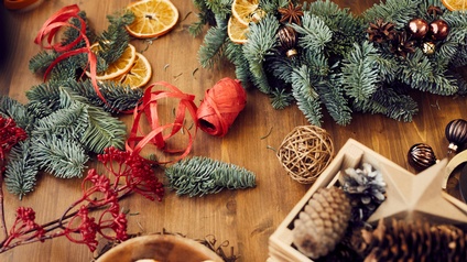 Auf einem Holztisch ist ein Adventkranz mit Dekoelementen wie Christbaumkugeln und getrockneten Orangenscheiben. Auf dem Tisch sind verschiedene Utensilien zum Basteln wie Tannenzweige oder Tannenzapfen