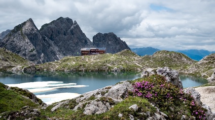 In der Mitte ist ein Bergsee. Um den See sind teilweise moos- und blumenbewachsene Steine. An einem Rand steht eine Hütte. Im Hintergrund ist eine massive Bergwand. Der Himmel ist wolkenverhangen.