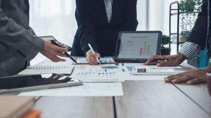 Um einen Tisch stehen drei Personen in Bürokleidung. Auf dem Tisch steht ein Tablet, auf dem Balkendiagramme sind. Darüber steht Immobilienbusiness. Auf dem Tisch liegen viele Zettel mit verschiedenen Diagrammen