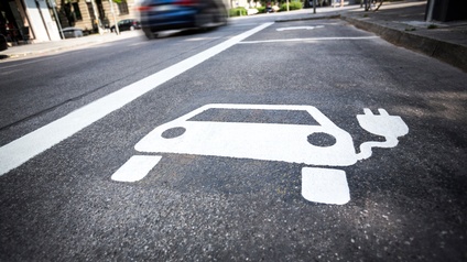 Auf einer asphaltierten Straße sieht man den Hinweis auf eine E-Lade-Station für ein Elektroauto als Bodenmarkierung in weißer Farbe. Daneben ist ein weitere Ladestation eingezeichnet. Links daneben ist eine Straße, auf der ein Auto fährt