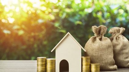 Auf einem Holzoberfläche stehen von links nach rechts zwei Münzstapel, ein Miniaturhaus, zwei Münzstapel und zwei Stoffsäcke. Im Hintergrund verschwommen sind grüne Pflanzen-