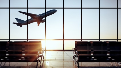 Ein moderner, leerer Flugsteig bei großen Glasfenstern, aus denen man bei Sonneneinstrahlung ein sich im Steigflug befindliches Flugzeug beobachten kann.