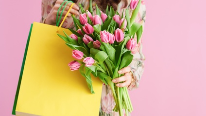 Person hält Einkaufstaschen in gelb und grün sowie einen Bund rosa Tulpen vor einem rosa Hintergrund