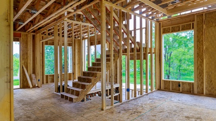 Innenaufnahme einer Baustelle eines Hauses. Wände und Boden bestehen noch aus einem hellen Material. In der Mitte führt eine Treppe nach oben. Im Hintergrund sind grüne Bäume und eine grüne Wiese