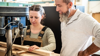 Zwei Personen unterschiedlichen Alters stehen in einer Tischlerei bei einer Bohrmaschine. Die jüngere Person blickt konzentriert auf ein Werkstück auf dem der Bohrer angesetzt ist. Die ältere Person schaut dabei zu. Beide wirken zufrieden.