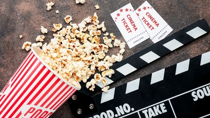 Popcornbehälter sowie Popcorn liegen auf einem Untergrund, daneben sind zwei Kinotickets sowie eine Filmklappe abgelegt