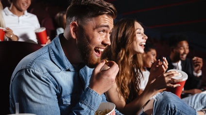 Mehrere Personen nebeneinander in Kinosaal sitzend schauen Richtung Leinwand und halten Getränke und Popcorn in Händen. Alle lachen