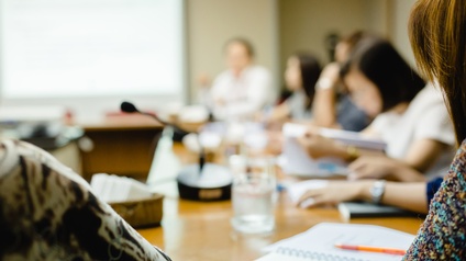 In einem Raum sitzen mehrere Personen um einen Tisch. Vor den Personen sind teilweise Unterlagen und Stifte. Auf dem Tisch steht ein Mikrofon. Im Hintergrund ist ein großer Bildschirm an der Wand