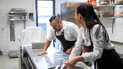 Zwei Personen in Schürzen stehen in einer Gastronomieküche und putzen jeweils mit Lappen die Oberflächen der Küche