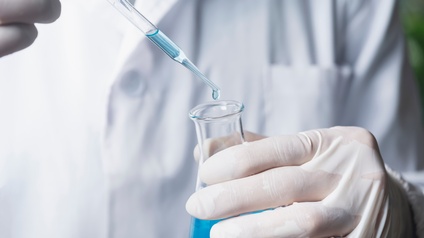 Hand mit Gummihandschuhen die aus Glaspipette blaue Flüssigkeit in Laborglas tropft