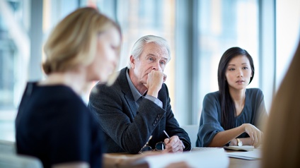 Drei Personen in Bürokleidung sitzen an einem Tisch. Im Fokus ist eine Person in einem dunklen Sakko sowie mit grauen Haaren. Auf dem Tisch liegen Papiere