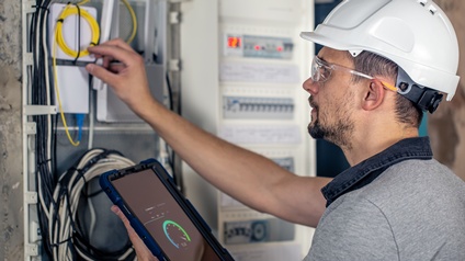 Seitliche Aufnahme einer Person mit Helm, Handschuhen und Schutzbrille, die neben einem Kasten mit verschiedenen Schaltern steht. In der linken Hand hält sie ein Tablet, auf der der Kasten mit den Schaltflächen abgebildet ist