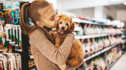 Ein Kind mit Zopf und braunem Oberteil hält mit beiden Armen einen kleinen, braune Hund, der das Maul geöffnet hat. Die Person hat ihre Lippen auf dem Kopf des Hundes und die Augen geschlossen. Im Hintergrund sind niedrige, lange Regale mit Tierartikeln.